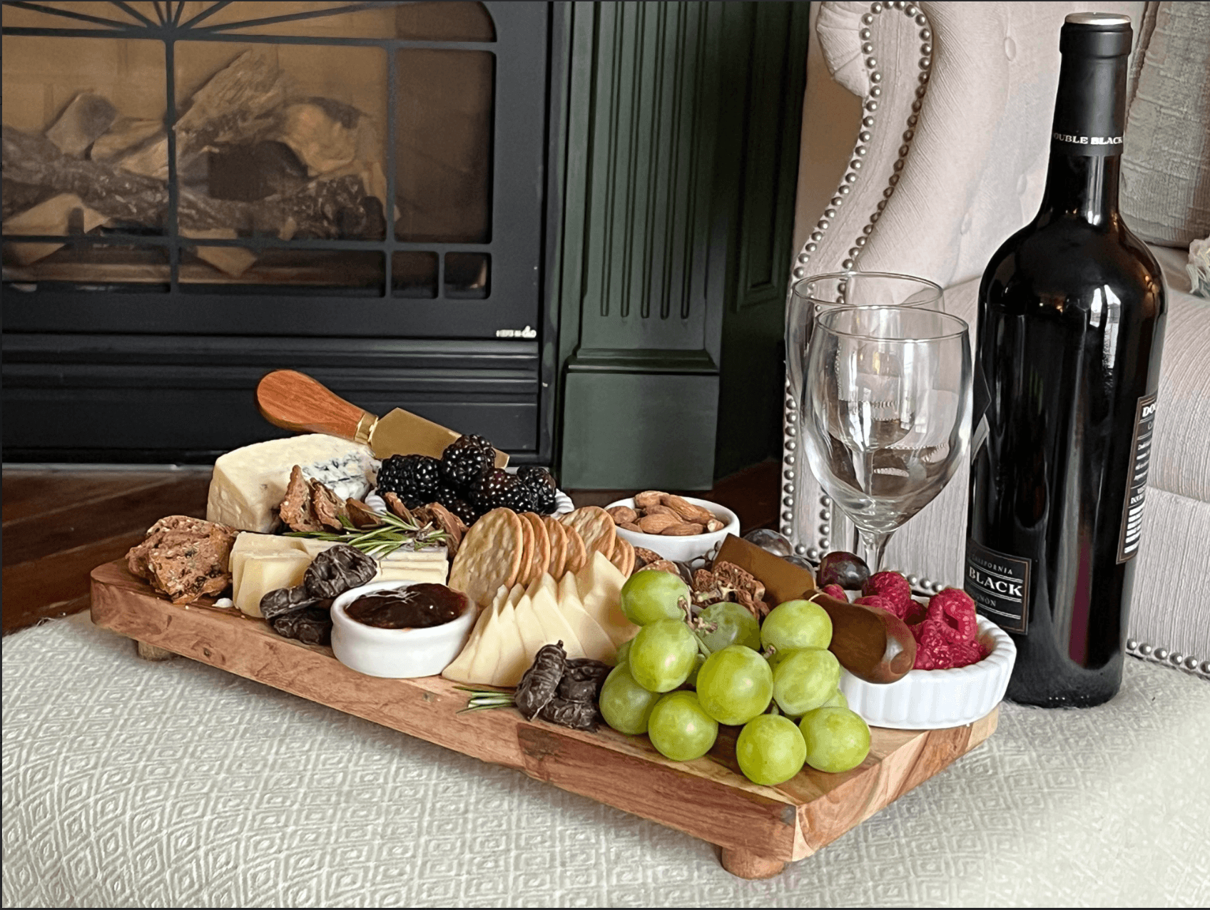 A wooden cheese board with assorted cheeses, fruits, nuts, and crackers, accompanied by a bottle of red wine and two glasses, set by a cozy fireplace.