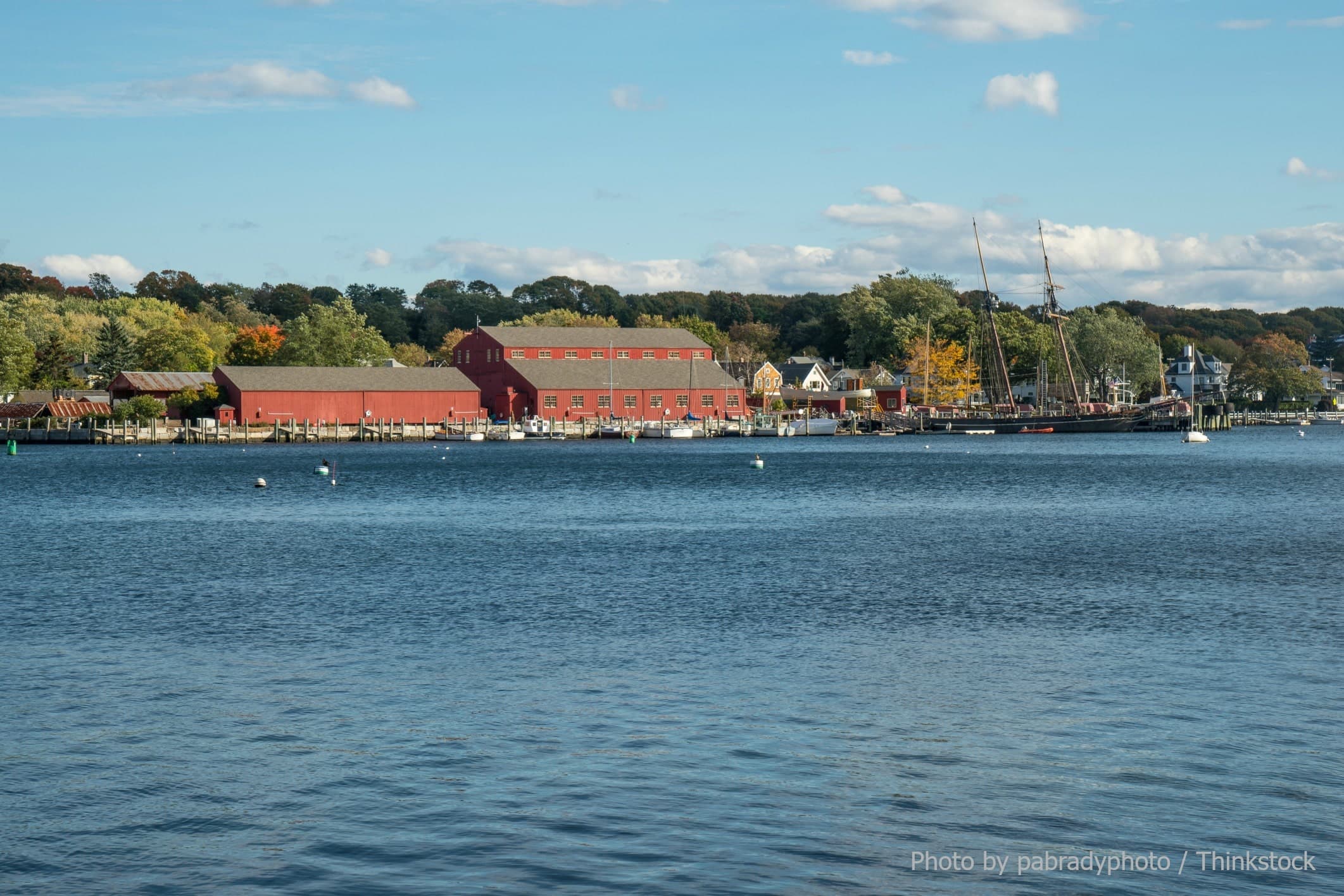A coastal scene featuring a red barn, boats, and trees in autumn colors alongside a calm waterway.