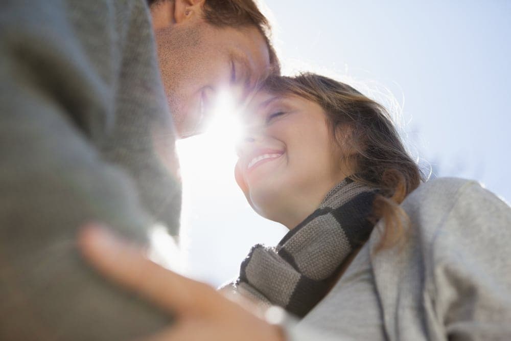 A couple smiles at each other, their foreheads touching in the sunlight.