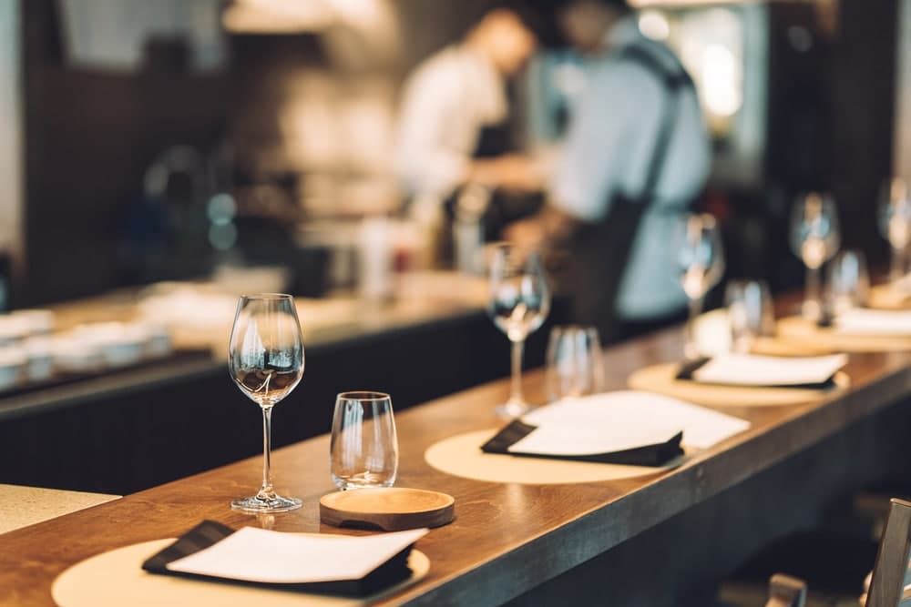 A dining table set with wine glasses and menus, with chefs working in the background.