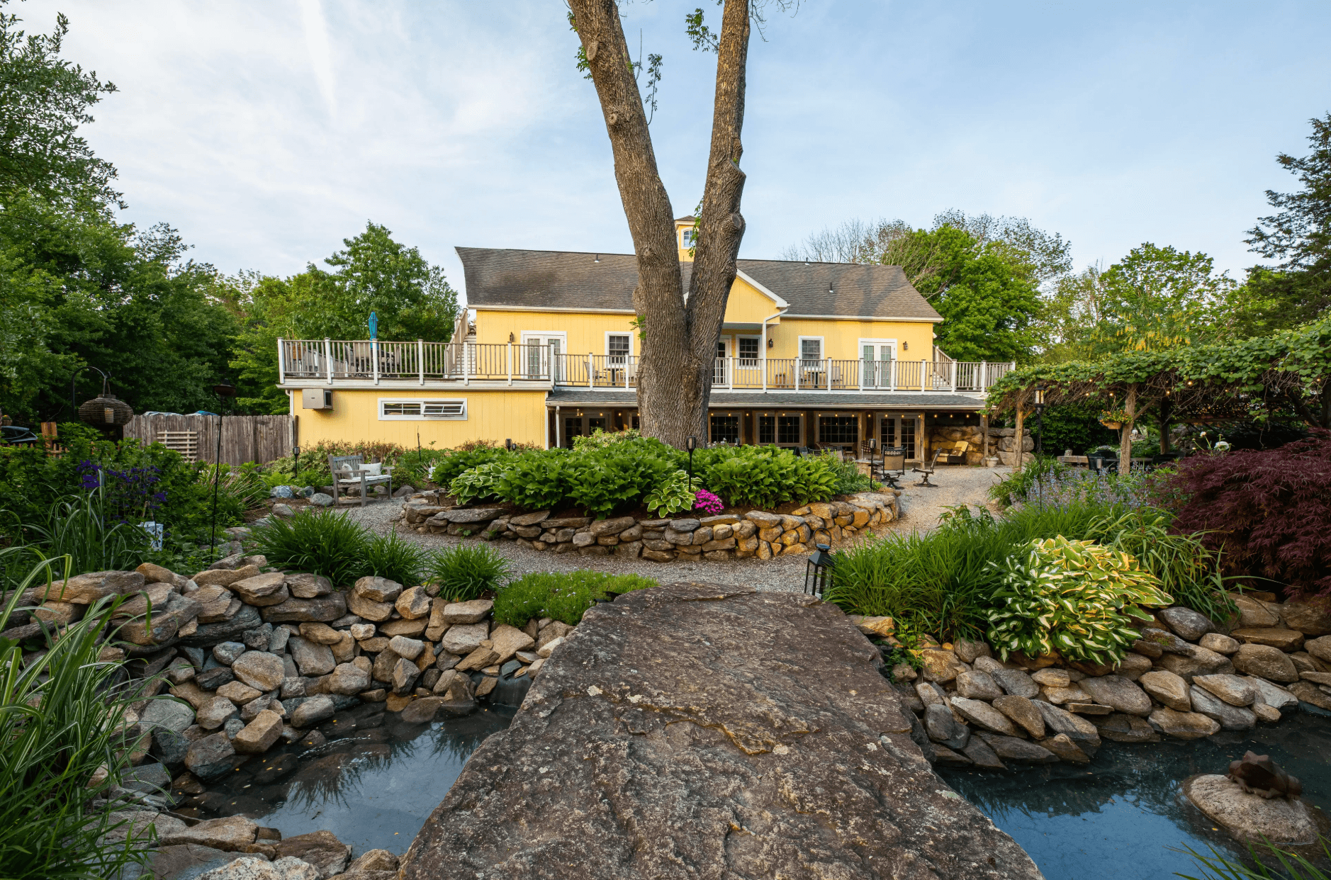 A yellow house with a deck surrounded by a landscaped garden and pond.