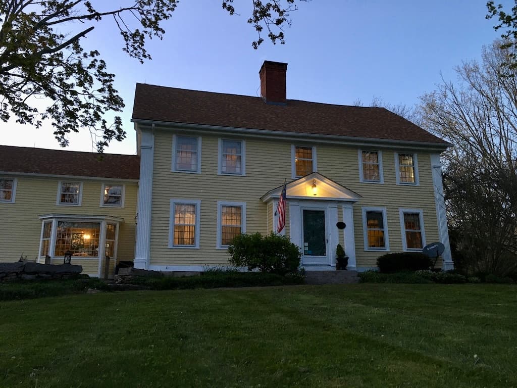A two-story yellow house with white trim, featuring a front porch and an American flag, is illuminated at dusk.
