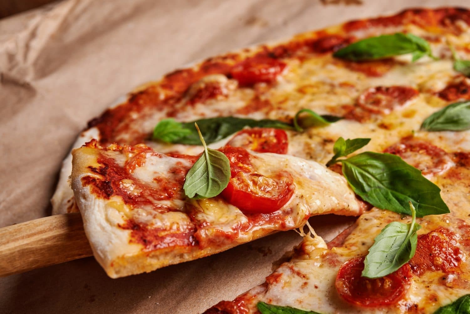 A slice of pizza with melted cheese, cherry tomatoes, and basil on a wooden spatula.