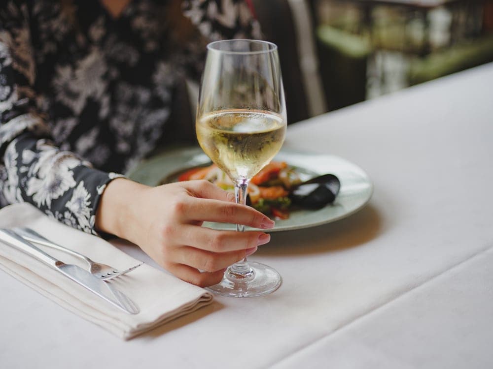A hand holding a glass of white wine next to a plate of food on a table.