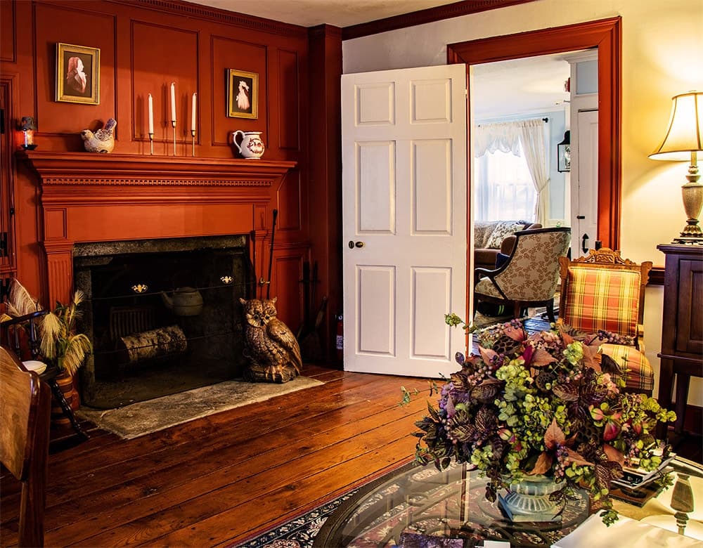 A cozy, warmly lit living room featuring an ornate fireplace, vintage decor, and a colorful floral arrangement on a glass table.