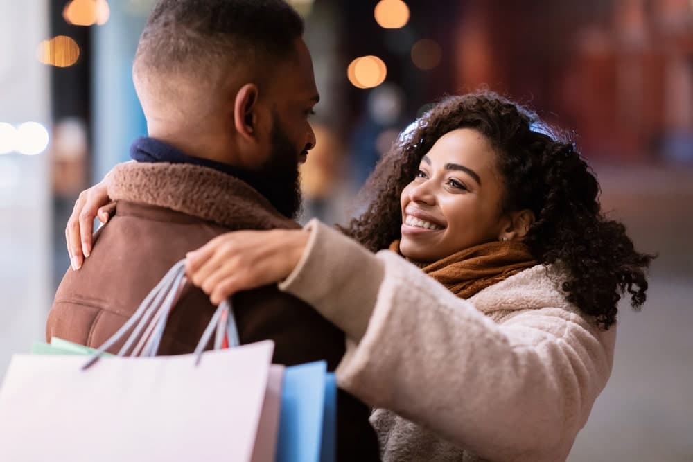 A couple shares a joyful moment while holding shopping bags in a festive, softly lit environment.