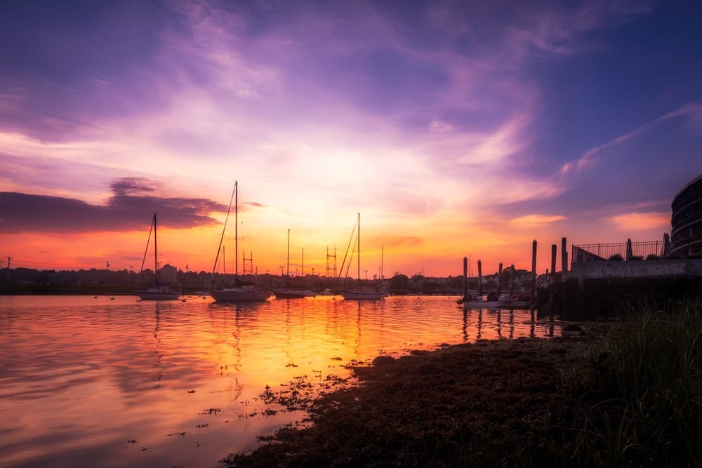 A serene sunset over a calm waterway with sailboats and silhouetted land in the background.