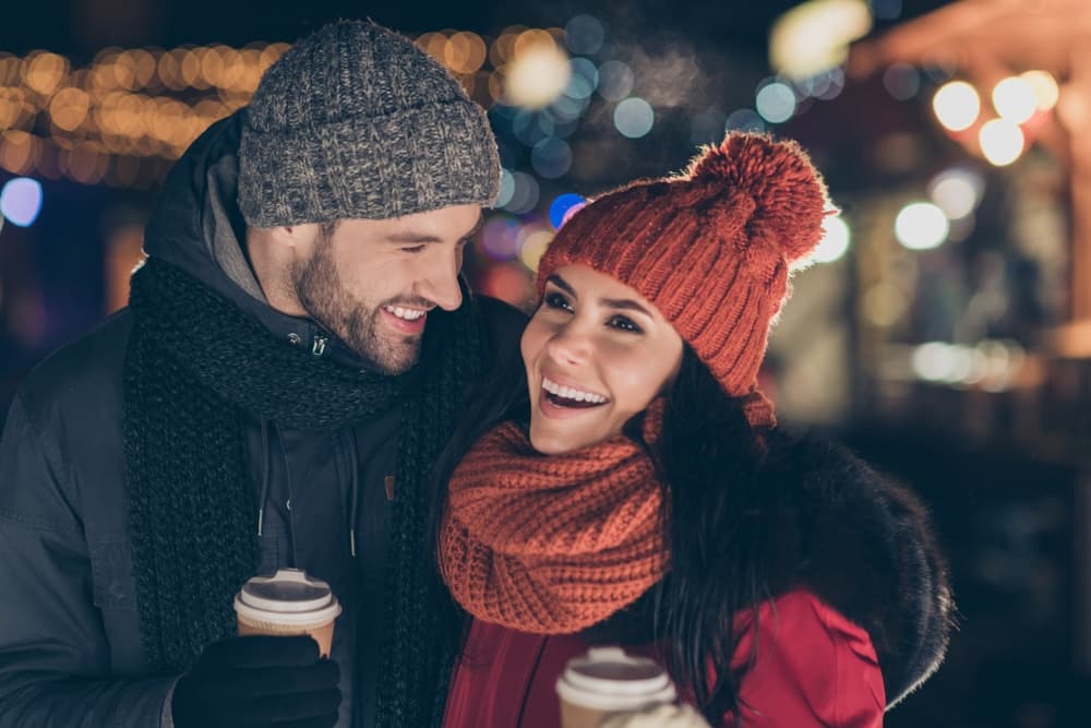 A smiling couple in winter attire share a joyful moment while holding coffee cups against a festive background.