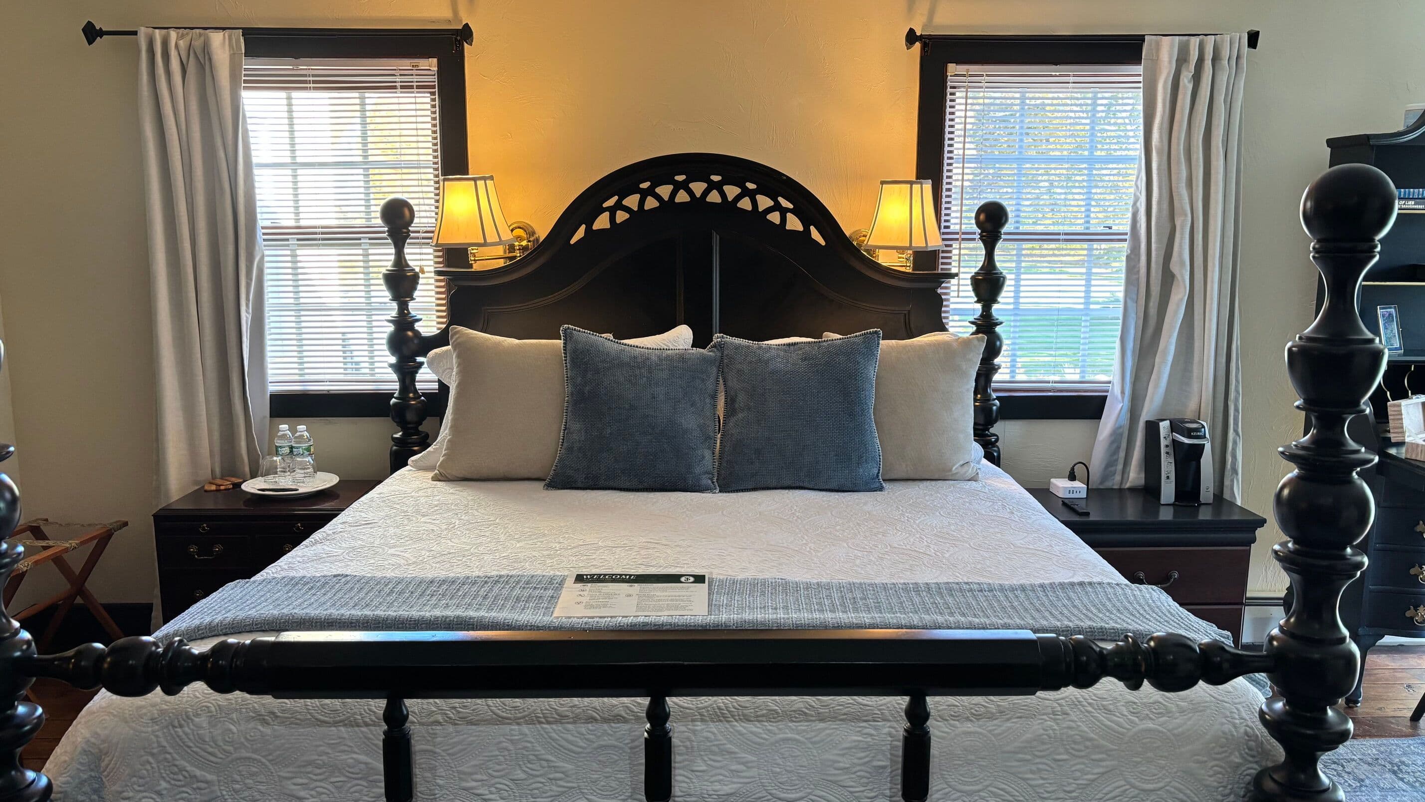 Master King Four Poster Bed with two white rectangular pillows and two blue square pillows. There are two windows on either side of the headboard.