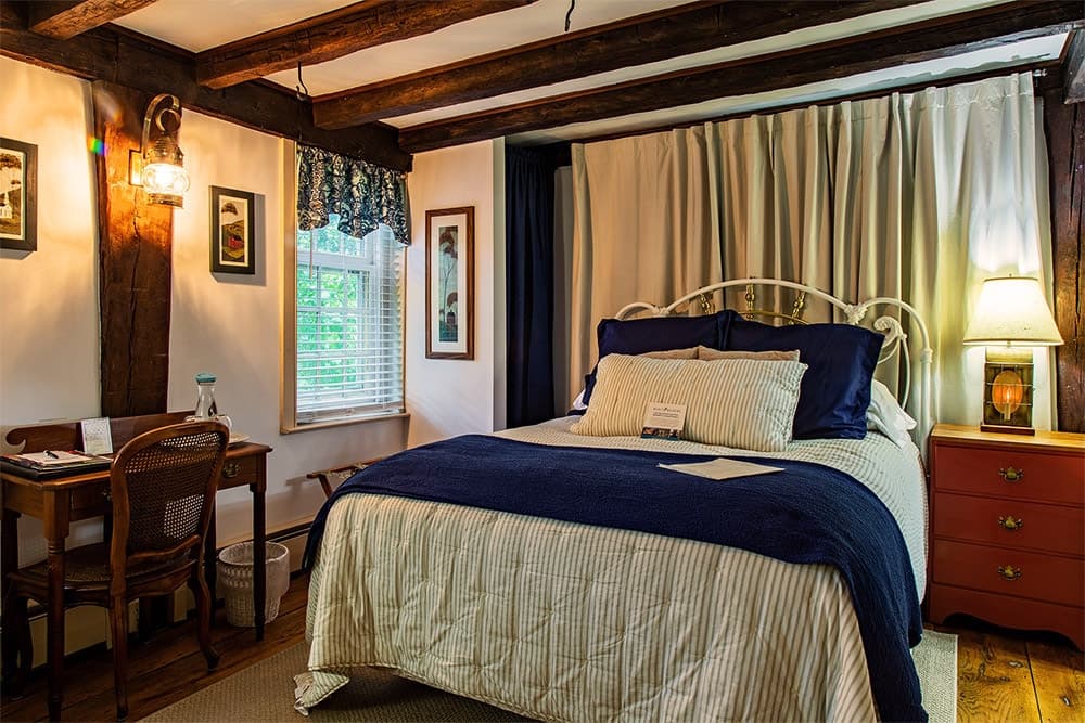 A cozy bedroom featuring a blue and white bedspread, wooden beams, a desk, and soft lighting.