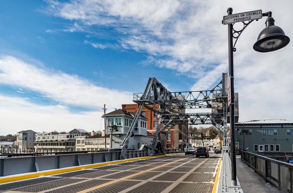 A drawbridge spans a road with buildings and a cloudy sky in the background.