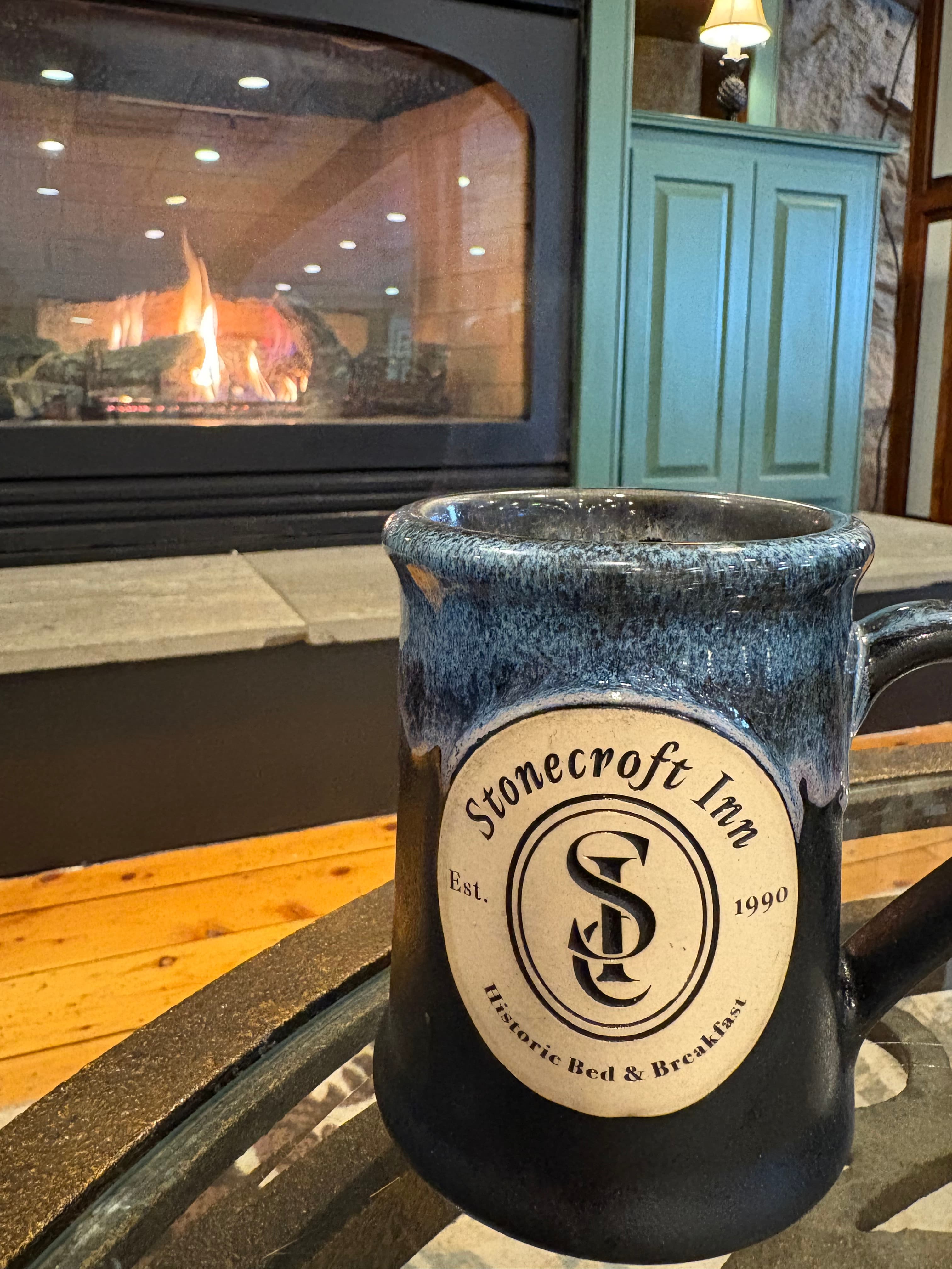 A mug with the "Stonecroft Inn" logo sits on a table in front of a cozy fireplace.