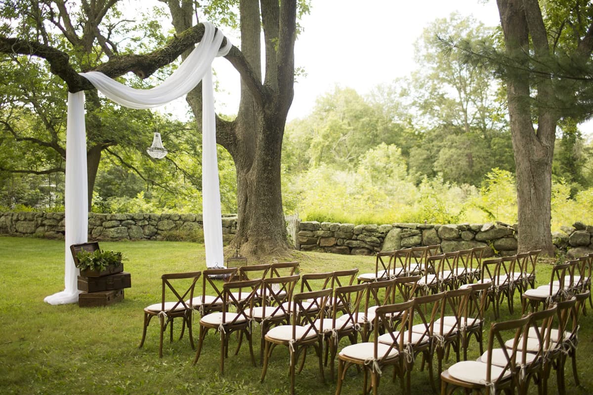 An outdoor wedding ceremony setup with empty chairs and a draped arch under trees.
