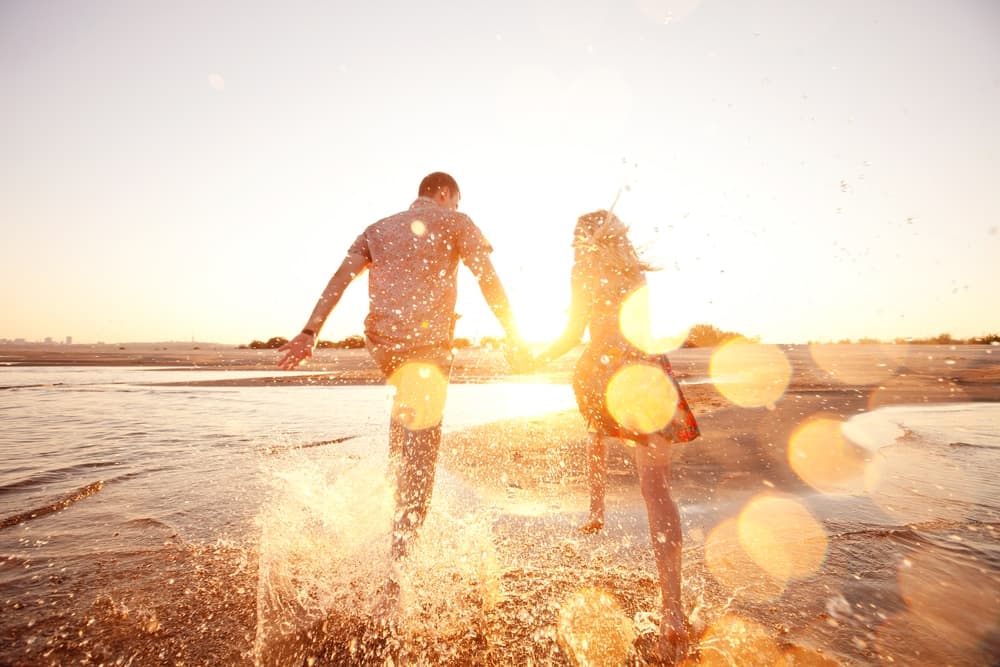A couple joyfully splashes through water at sunset, holding hands.