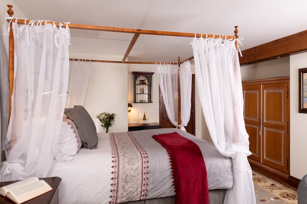 A cozy bedroom featuring a four-poster bed draped with white curtains, complemented by warm wood furnishings.