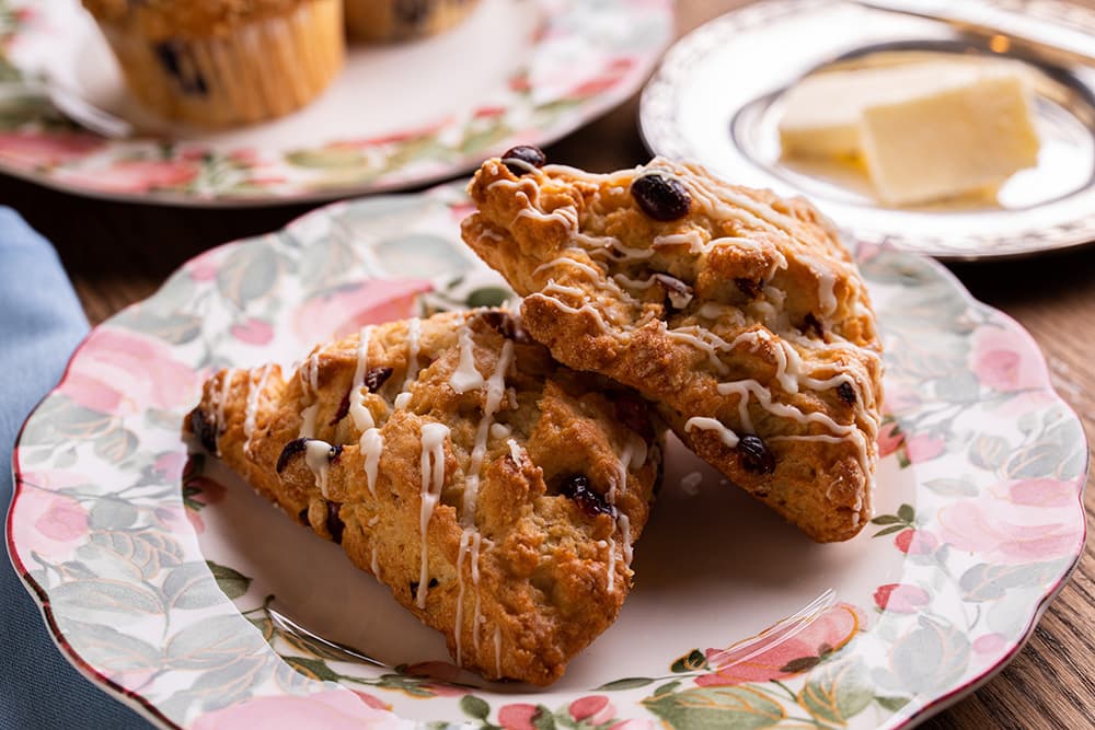 Two freshly baked scones with drizzled icing and dark raisins, served on a floral-patterned plate.