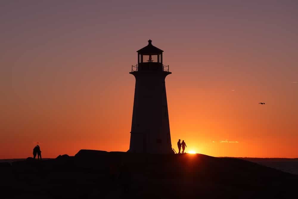 A silhouette of a lighthouse against a sunset, with people walking nearby.