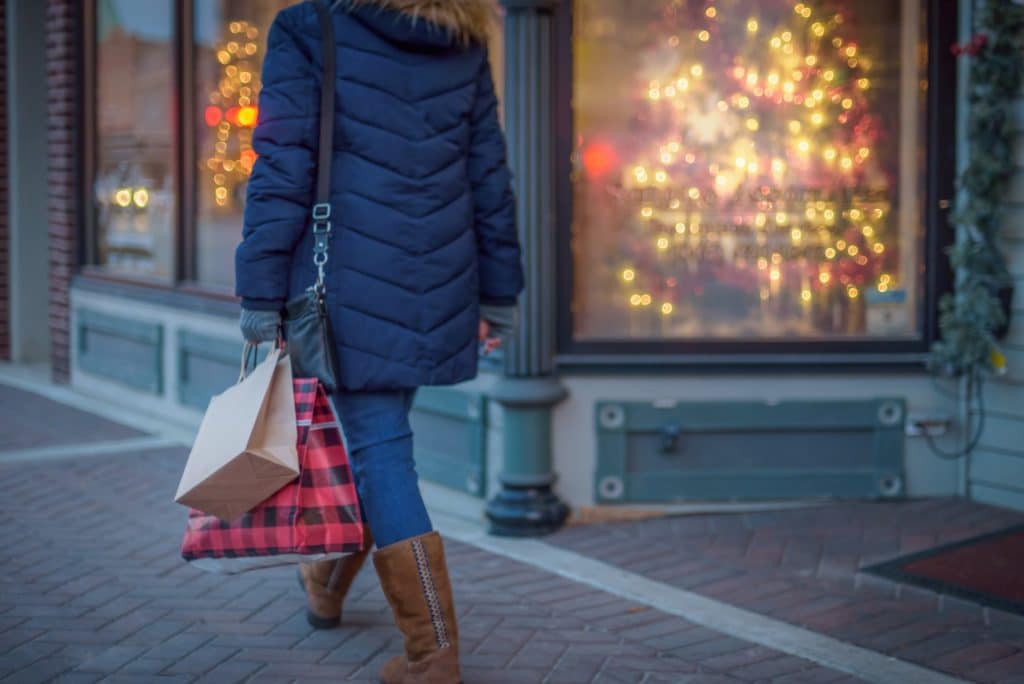 A person in a blue coat walks by a shop window adorned with festive lights, carrying two shopping bags.