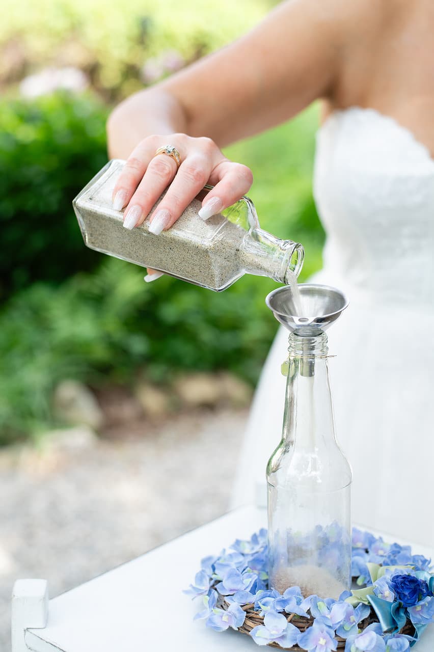 A person in a white dress pours sand from a glass bottle into a funnel resting on another glass bottle surrounded by blue flowers.