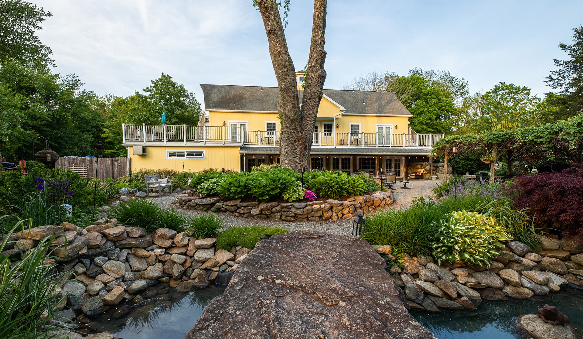 A yellow house with a balcony overlooks a landscaped garden and pond, featuring stone pathways and lush greenery.