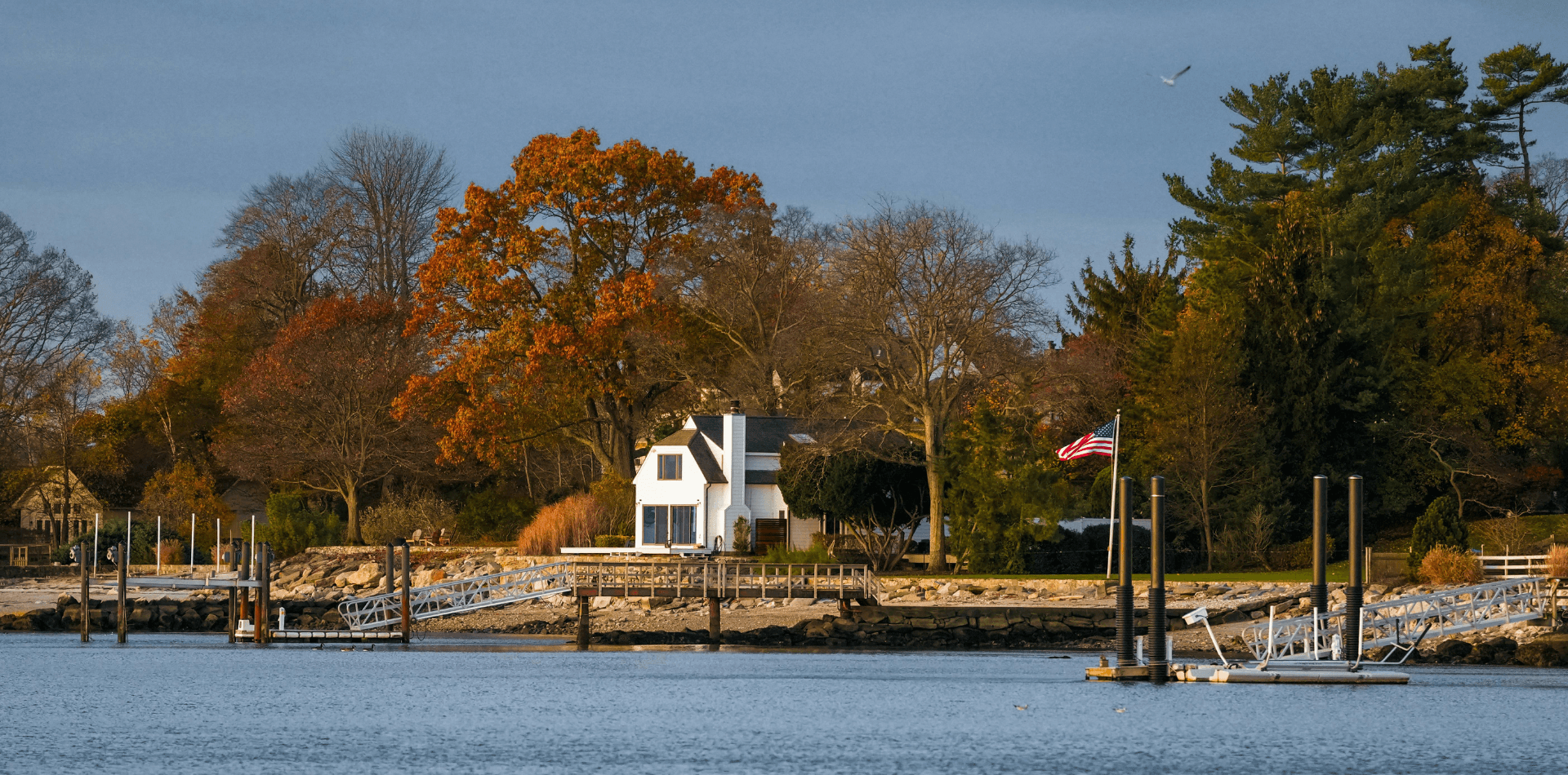 A tranquil waterfront scene featuring a white house surrounded by autumn trees and a dock with boats.