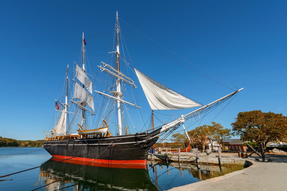 A tall ship with sails extended docked by a calm waterfront under a clear blue sky.