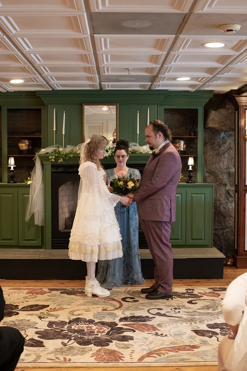 A bride and groom exchange vows during a wedding ceremony, led by an officiant in a cozy indoor setting.