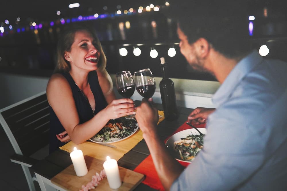 A couple enjoys a romantic dinner outdoors, toasting with glasses of wine against a backdrop of city lights.