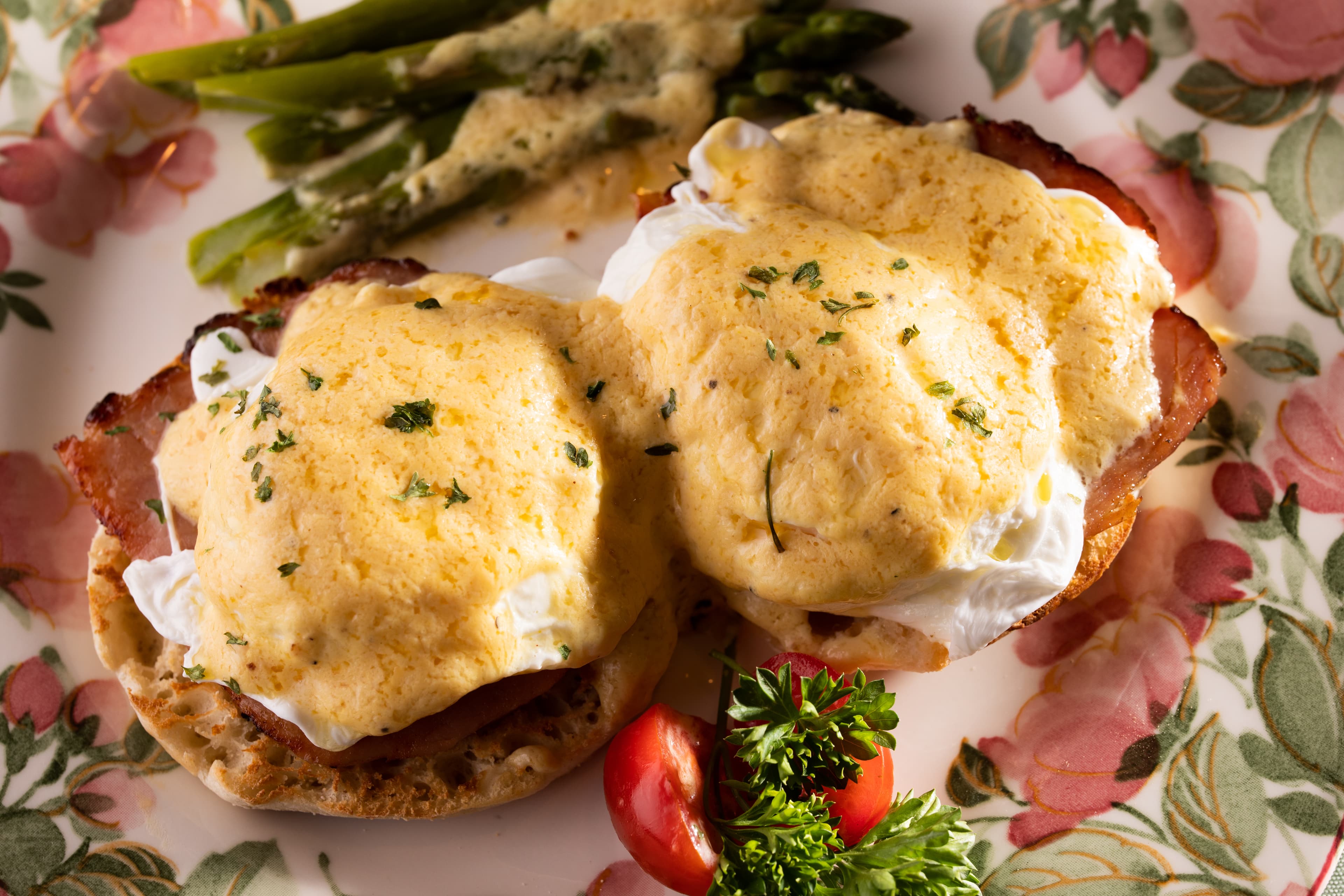 Two eggs Benedict on English muffins with ham, served with asparagus and cherry tomatoes on a floral plate.