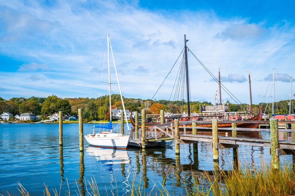 A sailboat is moored at a wooden dock beside a calm river, with trees and buildings in the background under a blue sky.
