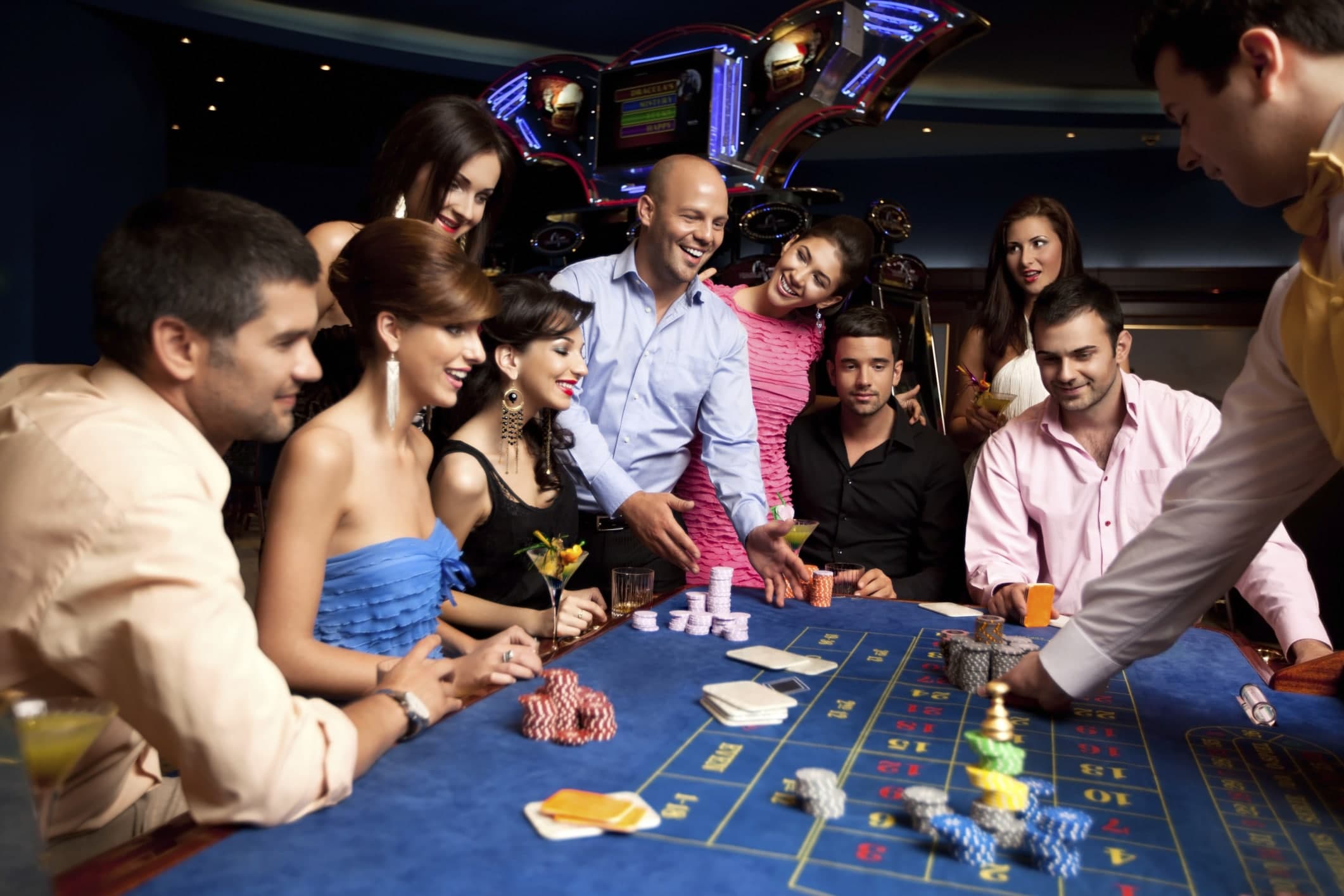 A group of well-dressed adults joyfully gather around a casino table with chips and drinks.