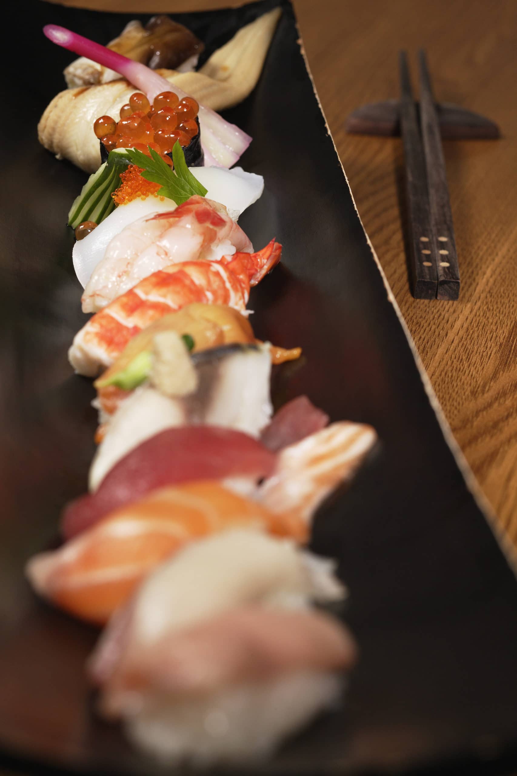 A black plate holds an assortment of sushi, featuring various types of fish and garnishes, with chopsticks nearby.