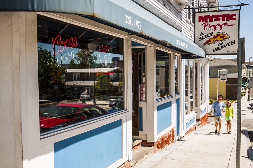 A couple walks by Mystic Pizza, a restaurant with a blue awning and a neon sign, in a sunny neighborhood.