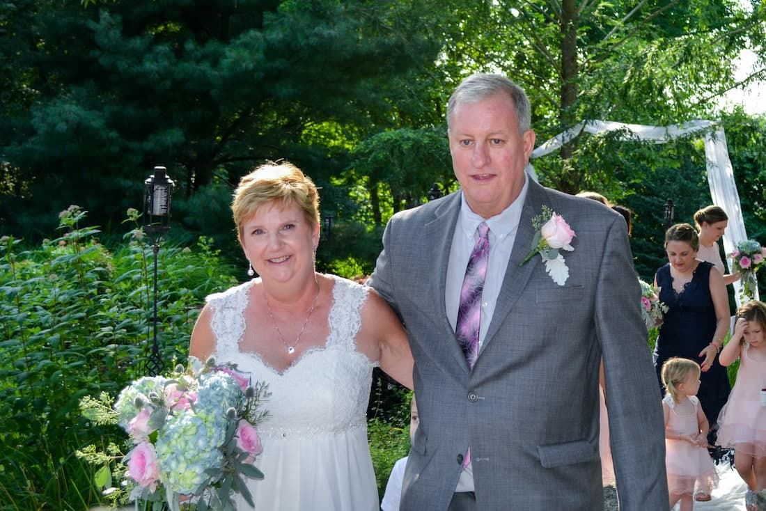 A woman in a white wedding dress and a man in a gray suit walk together, surrounded by greenery and wedding attendees in the background.