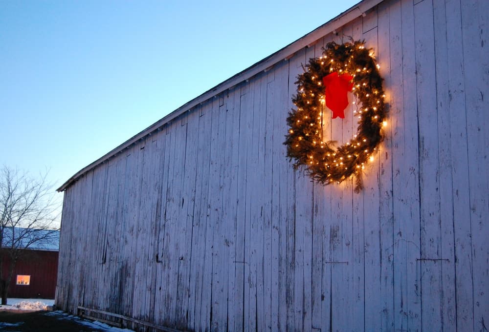 A decorated wreath with lights and a red bow hangs on a white barn against a twilight sky.