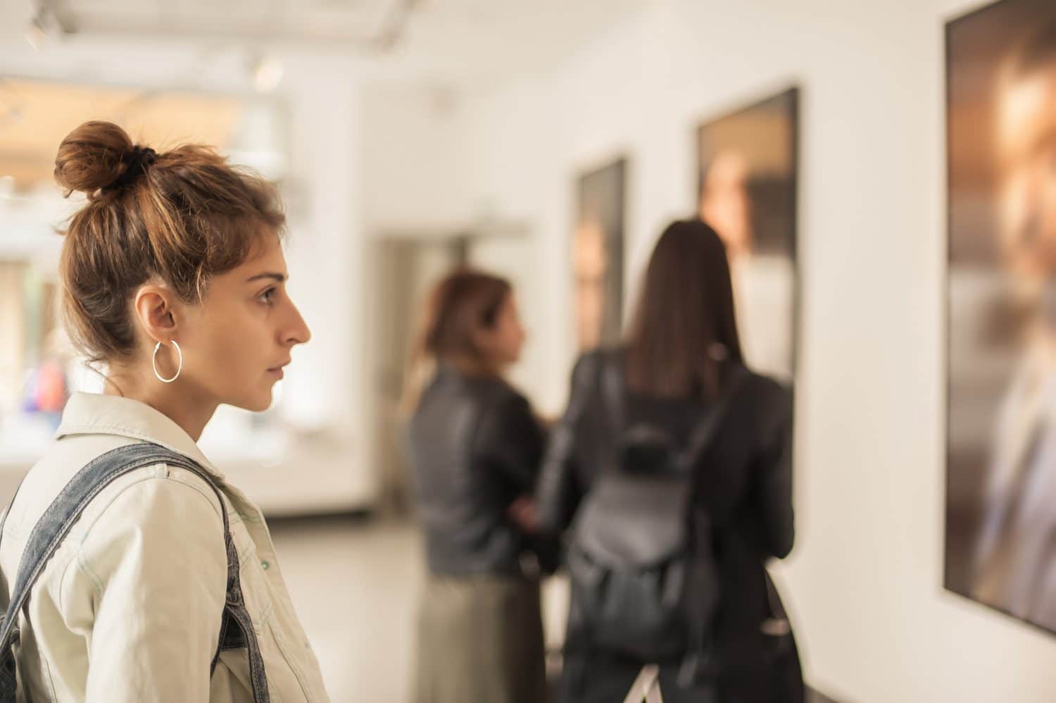 A young woman with a bun observes artwork in a gallery while two others stand behind her.
