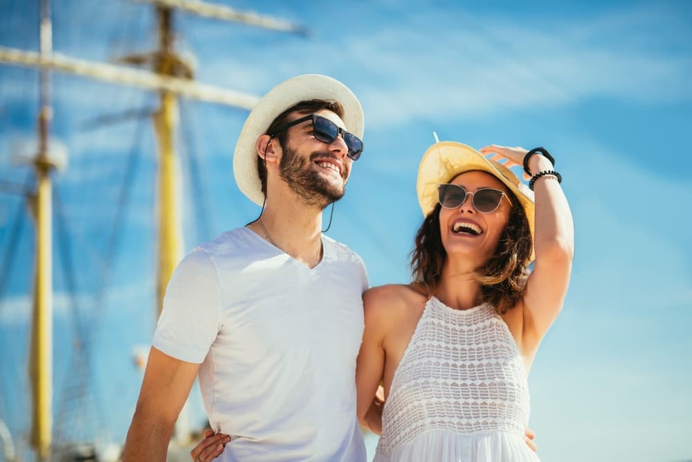 A smiling couple in summer attire poses joyfully at the beach with a ship in the background.
