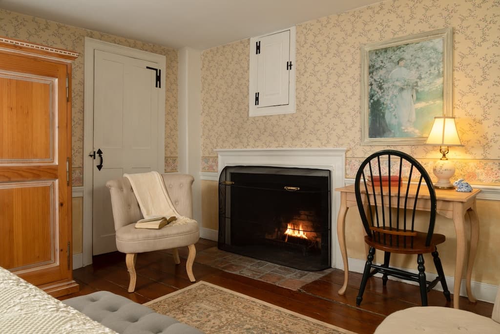 A cozy room featuring a fireplace, an armchair, a desk with a lamp, and decorative wallpaper.