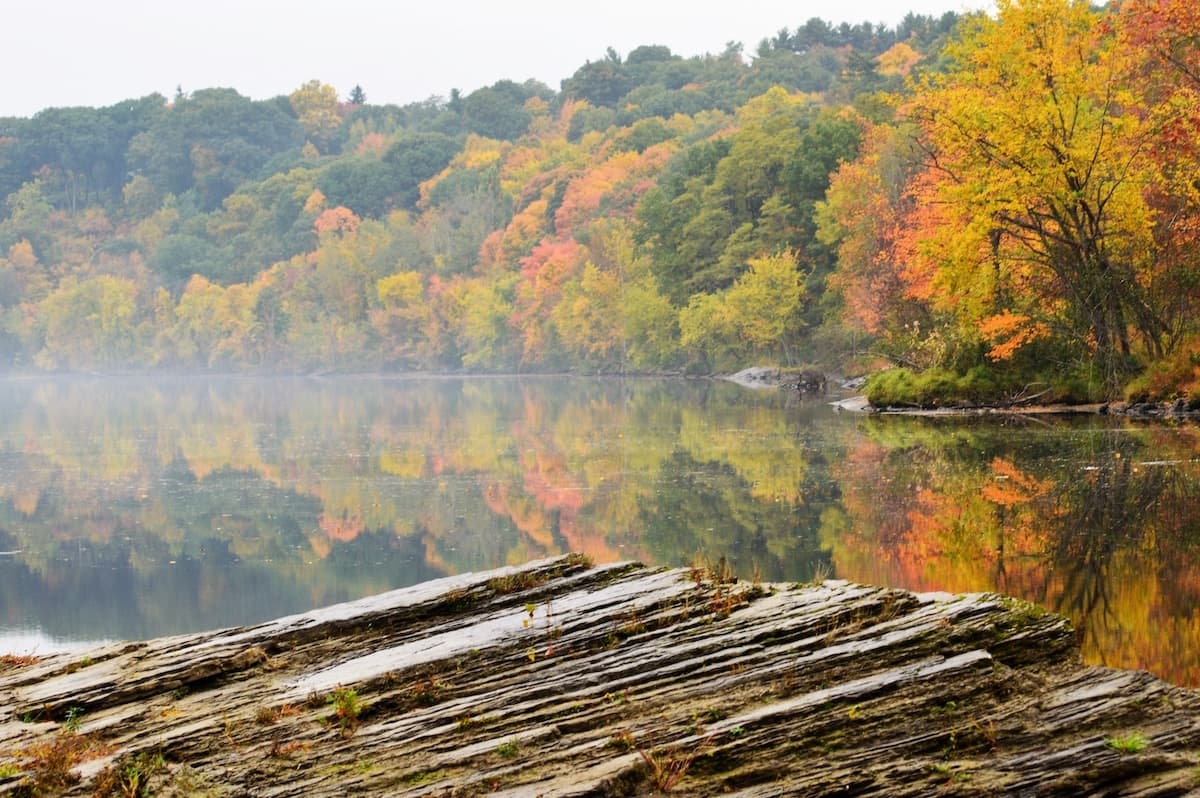 Autumn foliage reflects in a calm lake, with rocky foreground and misty hills in the background.