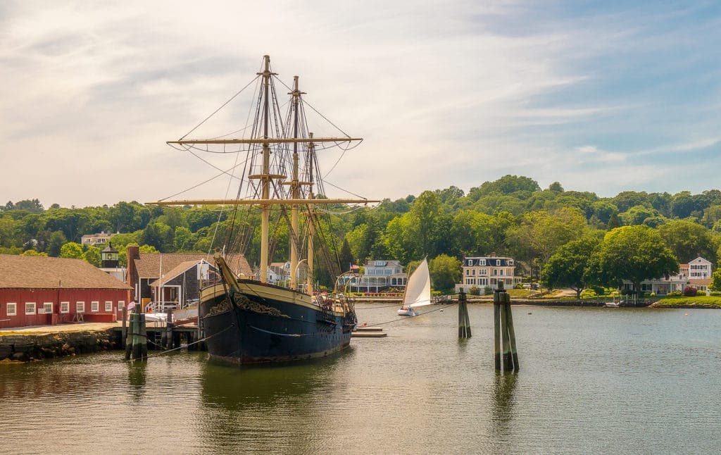 A tall ship docked in a serene harbor with trees and buildings in the background.