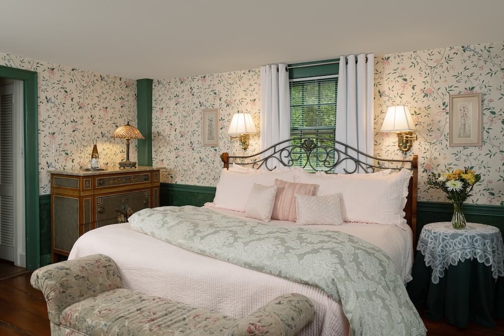 A cozy bedroom featuring floral wallpaper, a large bed with decorative pillows, and vintage furnishings.