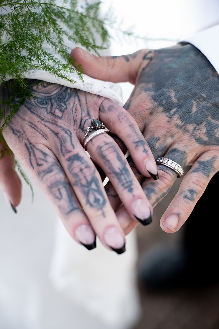 A close-up of two tattooed hands adorned with wedding rings, holding each other gently.