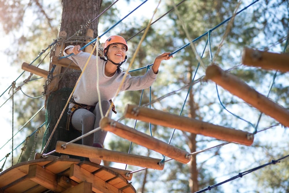 A woman navigates an elevated ropes course with a smile, wearing a safety harness and helmet.