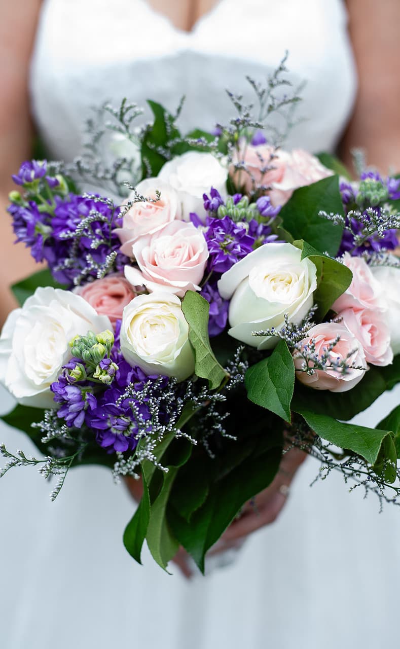 A bouquet of white and pink roses with purple flowers and greenery is held in front of a wedding dress.