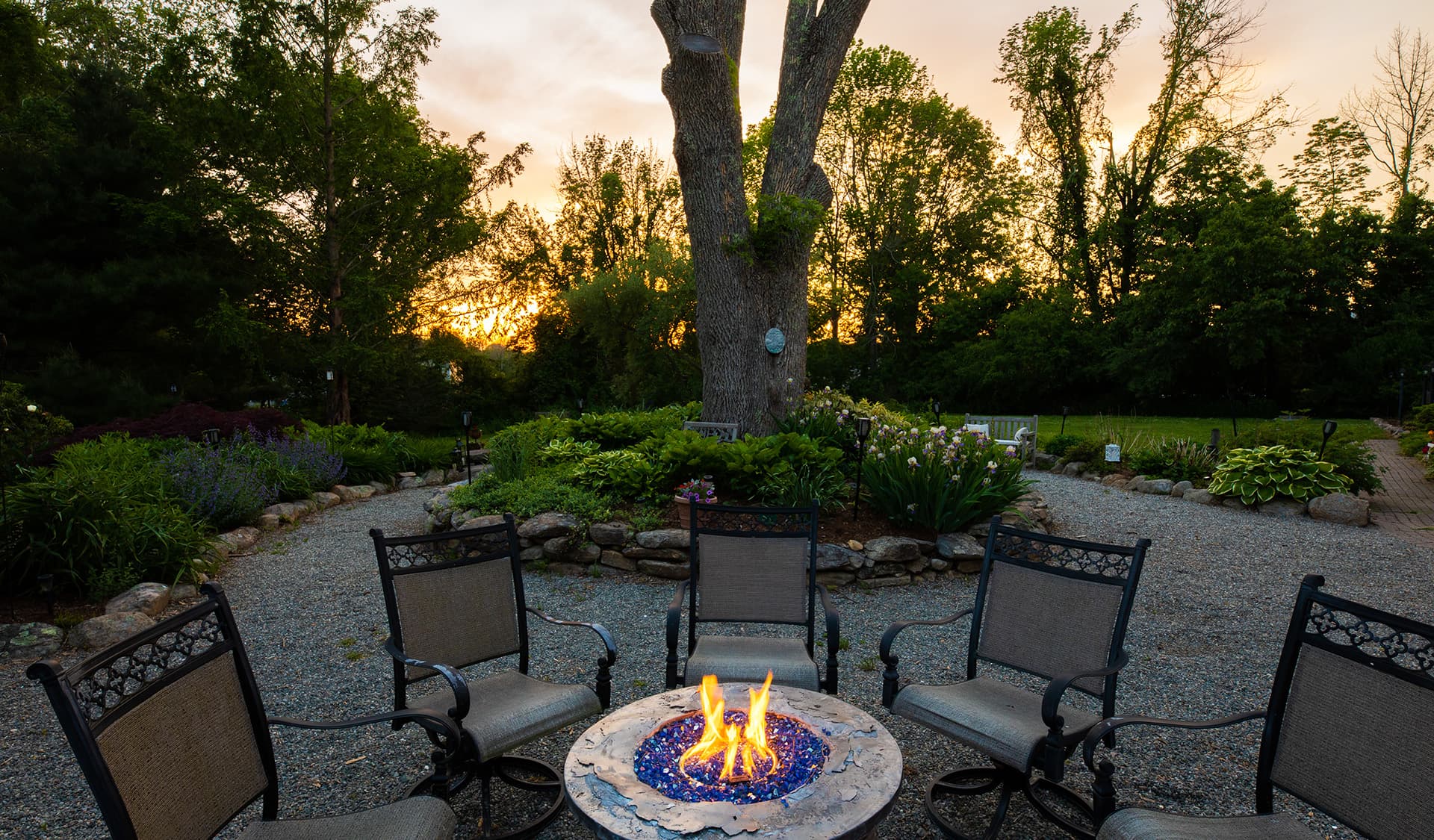 A cozy outdoor seating area with a fire pit surrounded by chairs, set against a sunset backdrop.