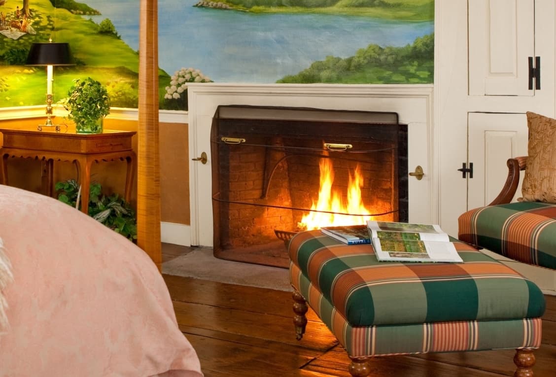 A cozy living space featuring a lit fireplace, a plaid ottoman, and a mural of a serene landscape.