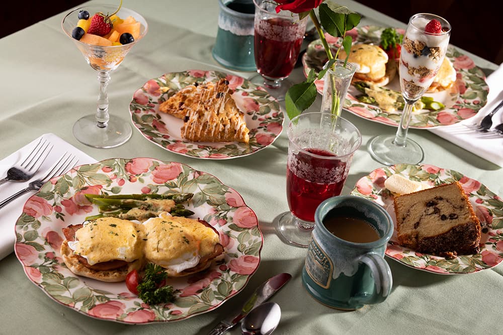 A beautifully arranged brunch table featuring various dishes on floral plates, including eggs Benedict, fruit, desserts, and beverages.