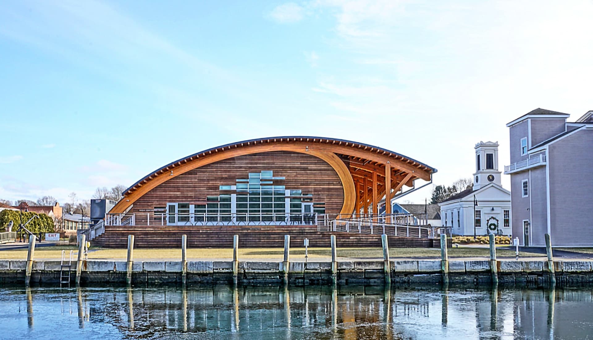 A modern wooden building with a curved roof reflected in calm water, alongside a traditional white structure.