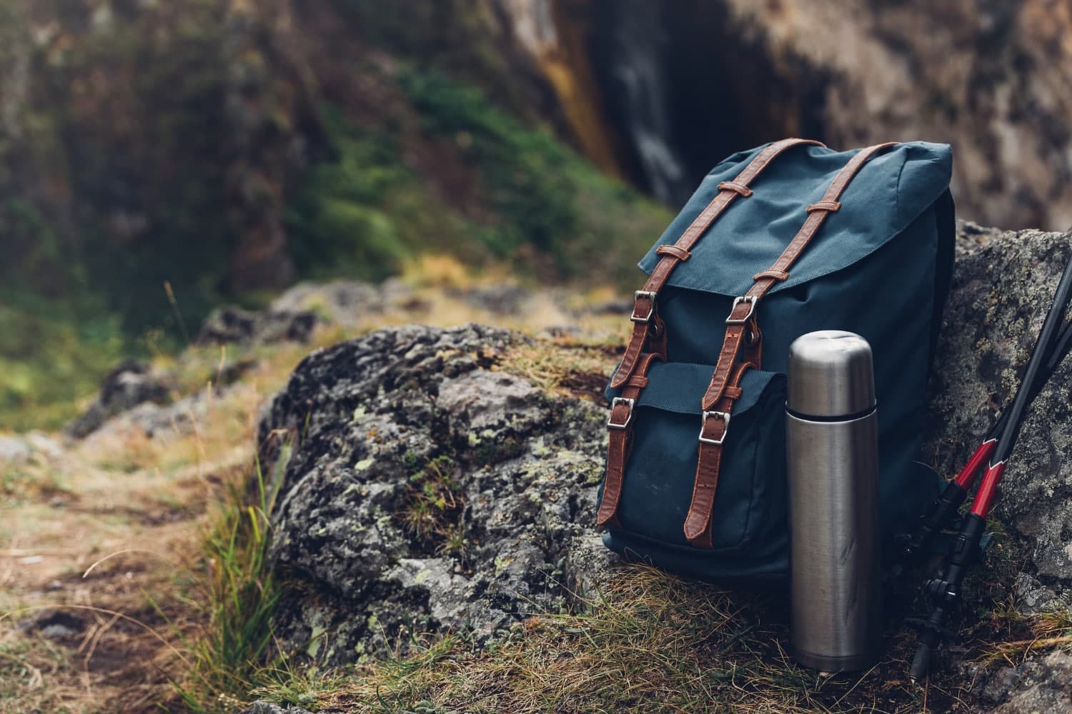 A blue backpack and a silver thermos sit on a rocky surface in a natural setting. A blue backpack and a silver thermos sit on a rocky surface in a natural setting.