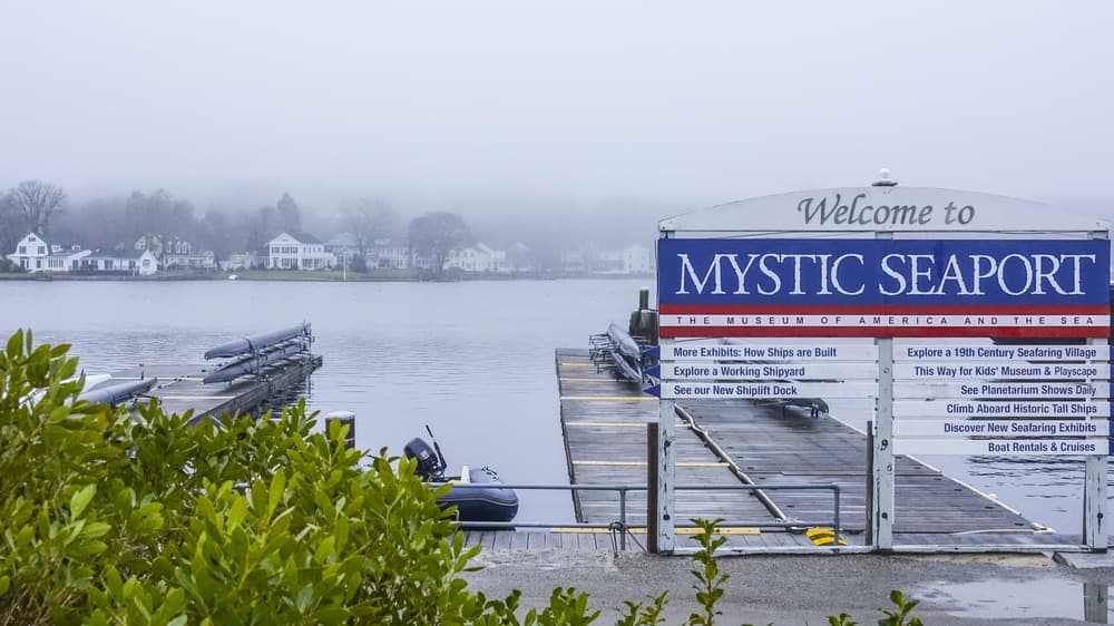 A foggy view of the Mystic Seaport sign beside a calm waterfront and dock.